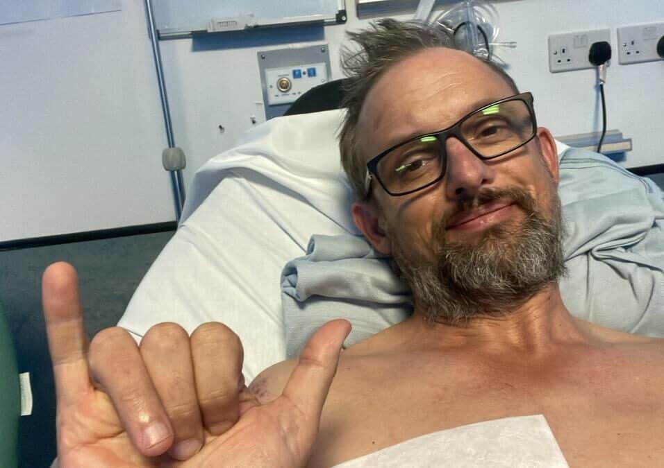 A man in glasses giving a thumbs-up while lying in hospital bed after treatment, smiling confidently.