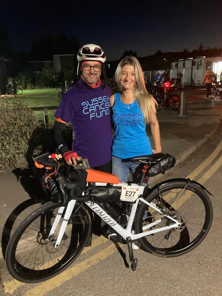 A man and woman in branded cycling gear with a mountain bike at night.